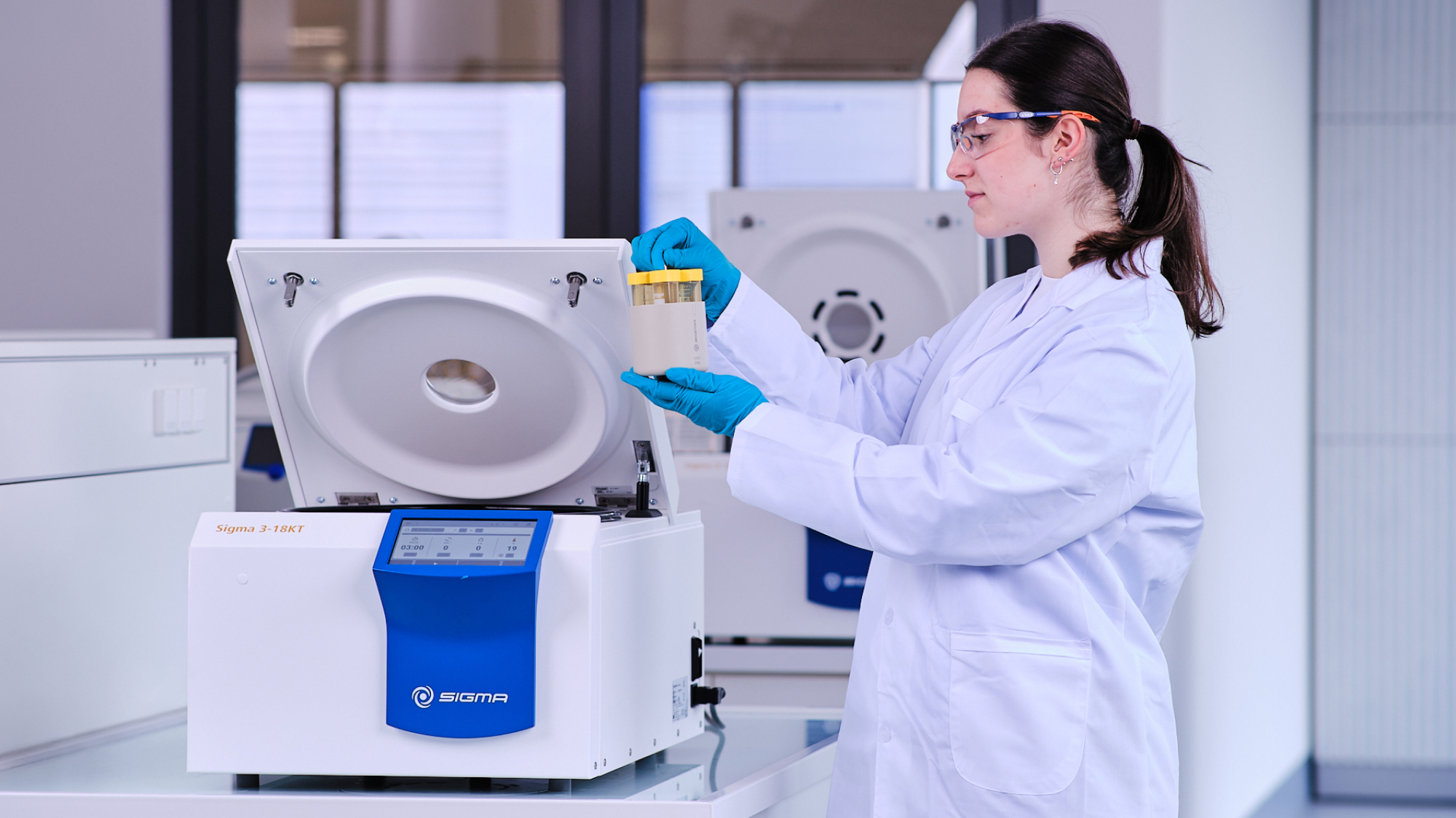 Lab technician in a white coat, safety glasses and blue gloves loading sample tubes into the open Sigma 3-18KT refrigerated centrifuge